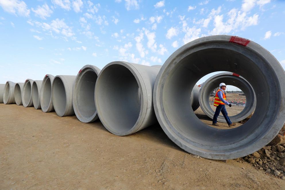 A construction worker wearing a hard hat and safety vest walks through the opening of a large concrete pipe, which is part of a long row of pipes lined up on a dirt construction site.
